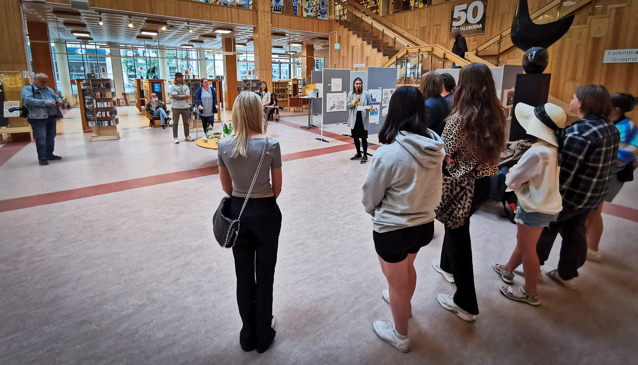 Bilden visar en grupp ungdomar som lyssnar på en kvinna med mikrofon på stadsbiblioteket i Falun