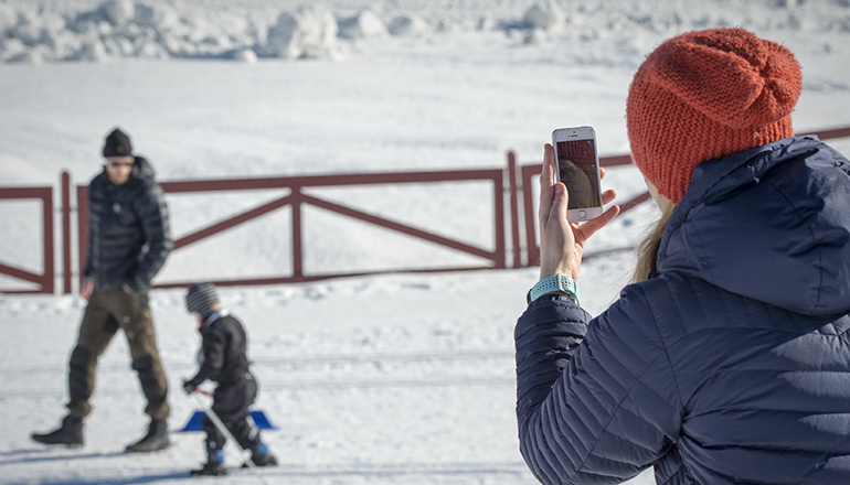 Kvinna med mobil filmar barn och man som åker skidor